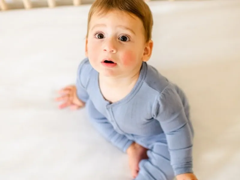 Baby in blue pyjamas sitting awake in an empty white crib, looking up, for baby sleep help in Calgary