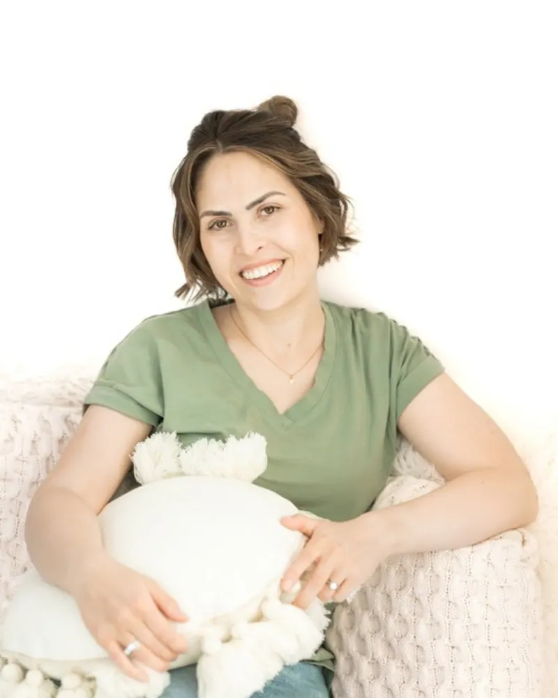 Jenna Phan, mom and Calgary pediatric sleep consultant, smiling in a green tee and holding a cushion on a cream chair