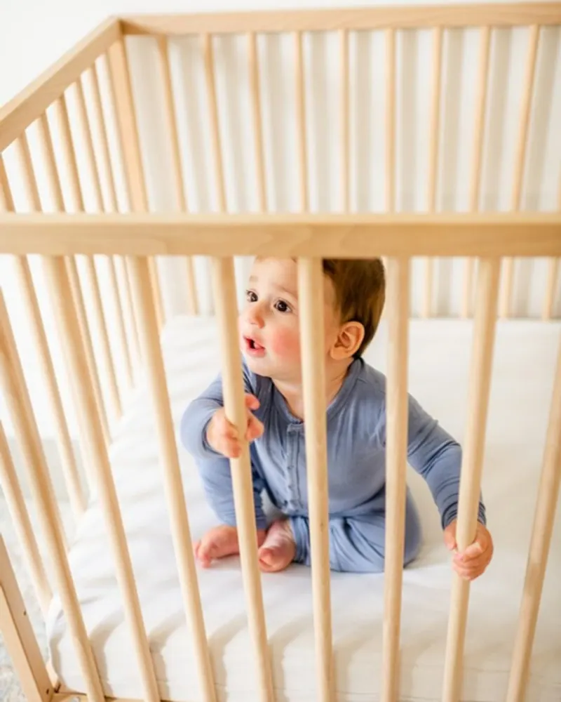 Baby in blue pyjamas standing at the rails of an empty wooden crib, ready for gentle baby sleep training