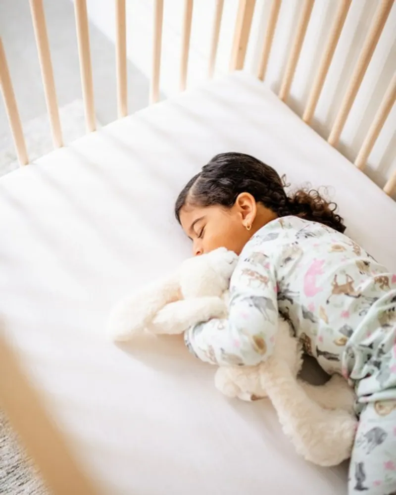 Toddler girl in animal-print pyjamas asleep on her side in a wooden crib, hugging a cream stuffed bunny